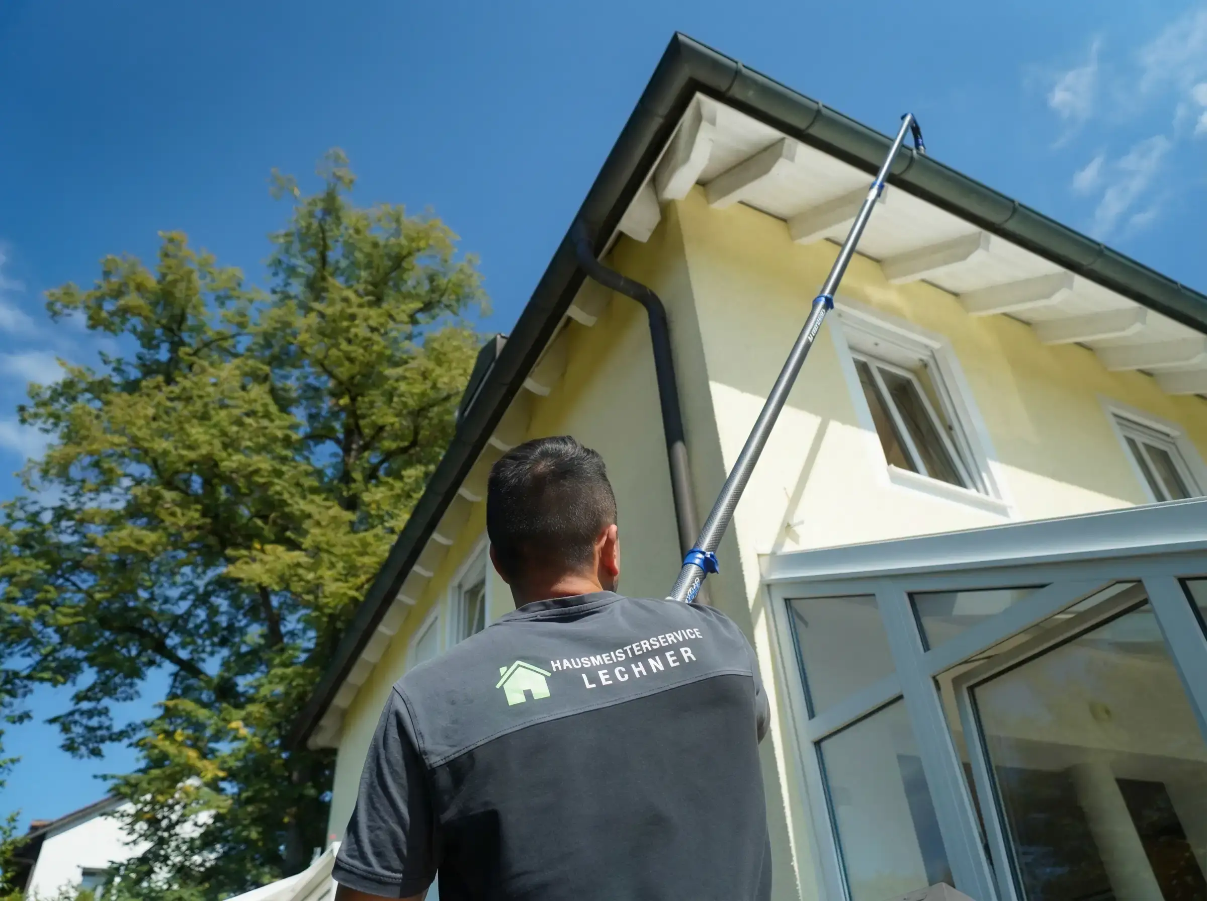 Hausmeister in Uniform reinigt Fenster eines gelben Hauses mit Teleskopstab unter blauem Himmel.