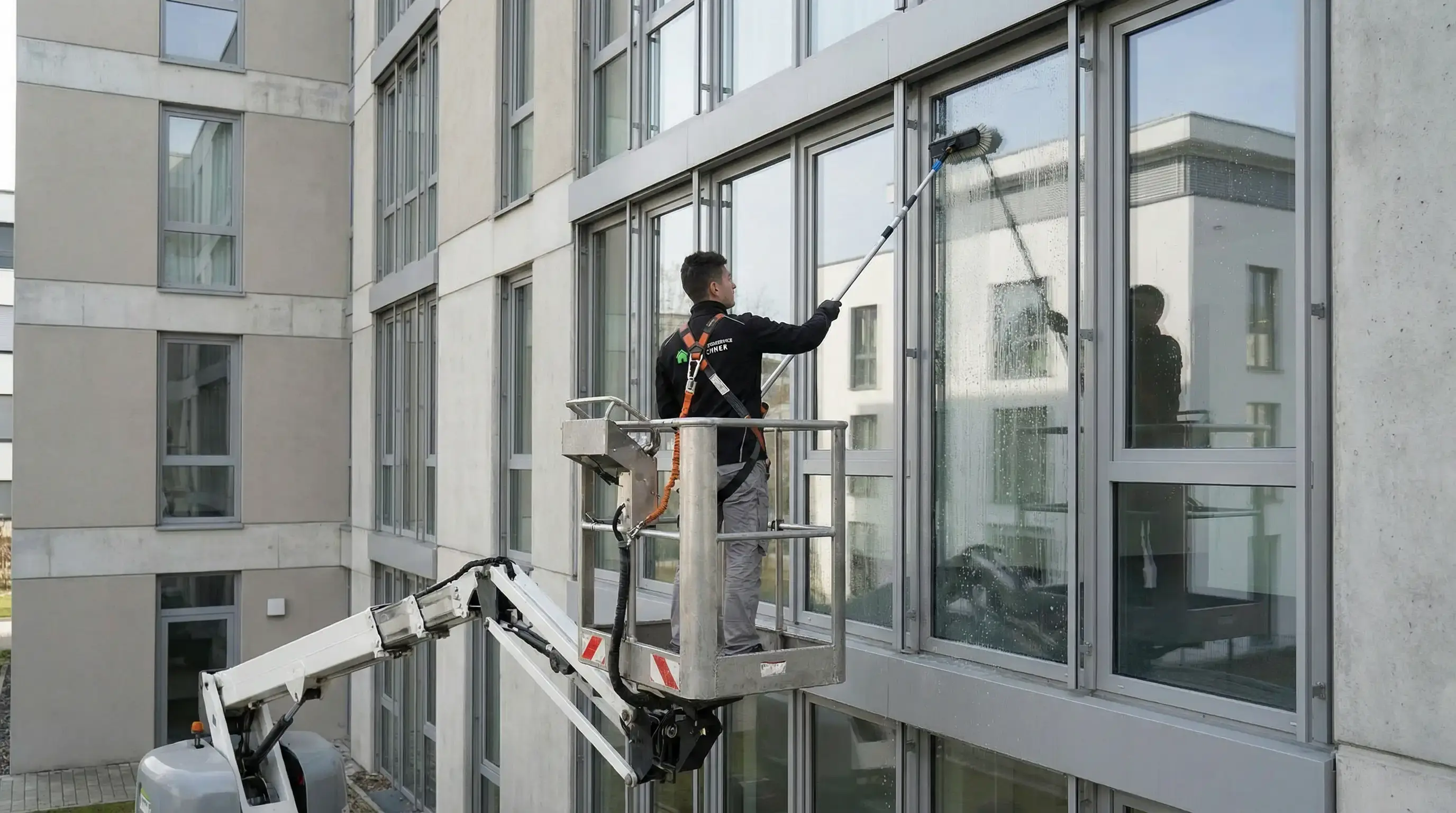 Mann reinigt mit einer Bürste an einem Teleskopstiel hohe Fenster an einem modernen Gebäude von einer Hebebühne aus.