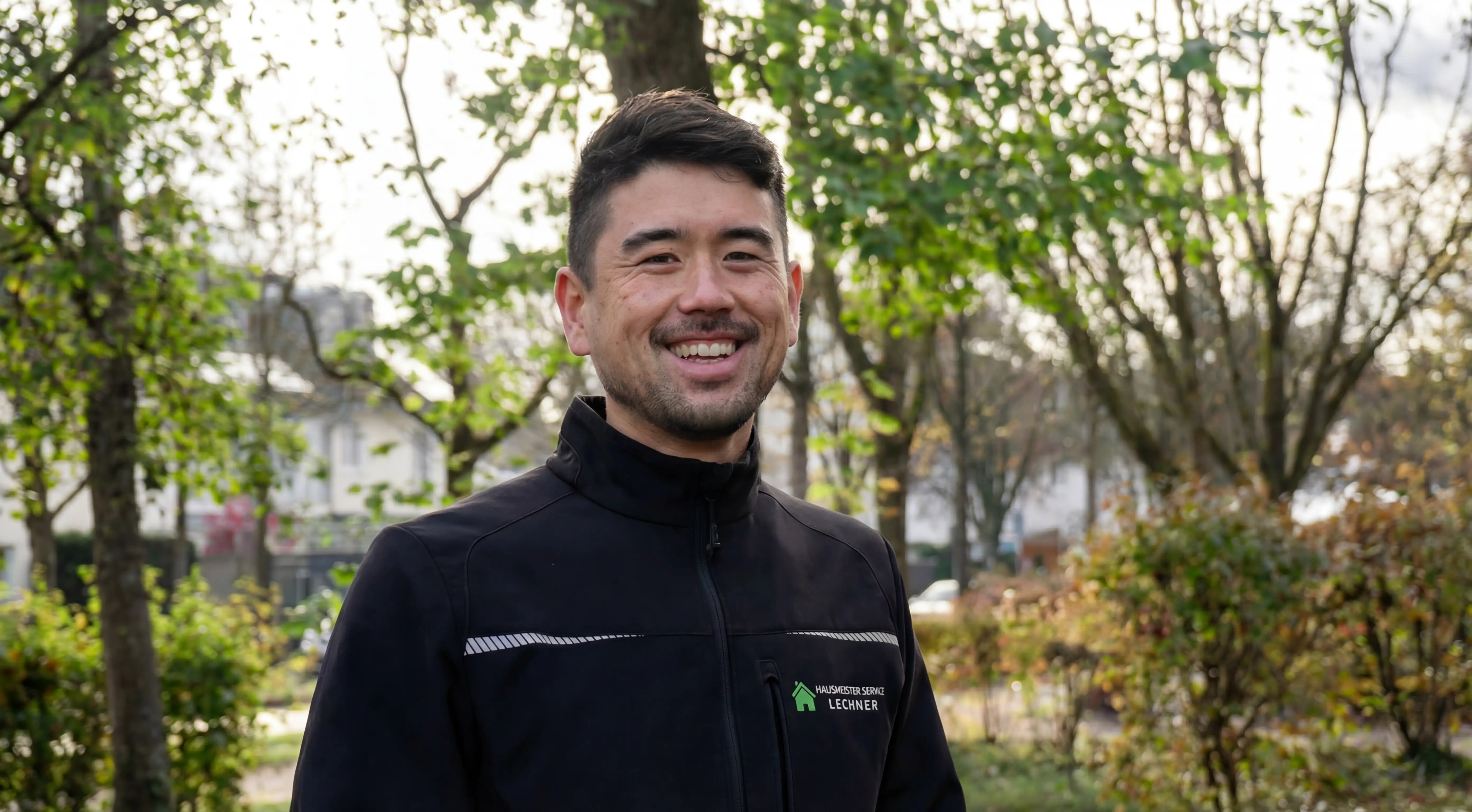 Lächelnder Mann in schwarzer Jacke mit Hausmeister Service Lechner Logo, stehend in einem Park mit Bäumen.