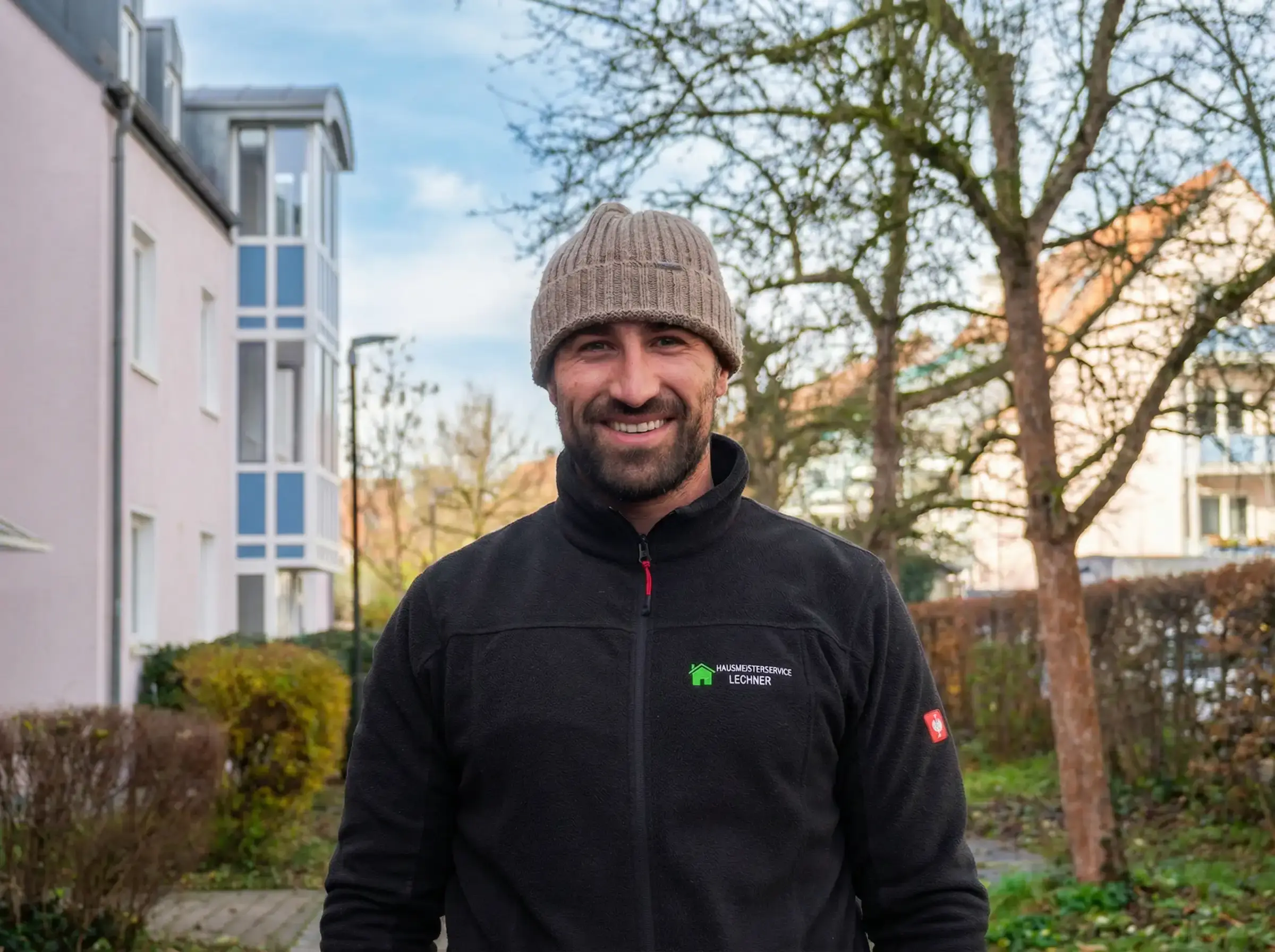 Mann mit brauner Strickmütze und schwarzer Jacke mit Hausmeisterservice Lechner Logo lächelt im Freien vor Wohngebäuden.