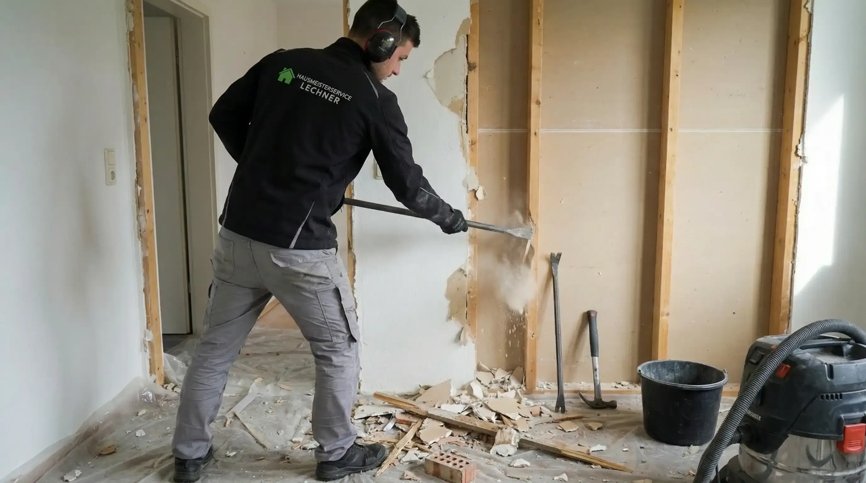 Ein Mann mit werkstatttauglicher Kleidung und Gehörschutz entfernt mit einem Brecheisen eine Wandverkleidung in einem Raum mit Bau- und Abbruchmaterial am Boden.