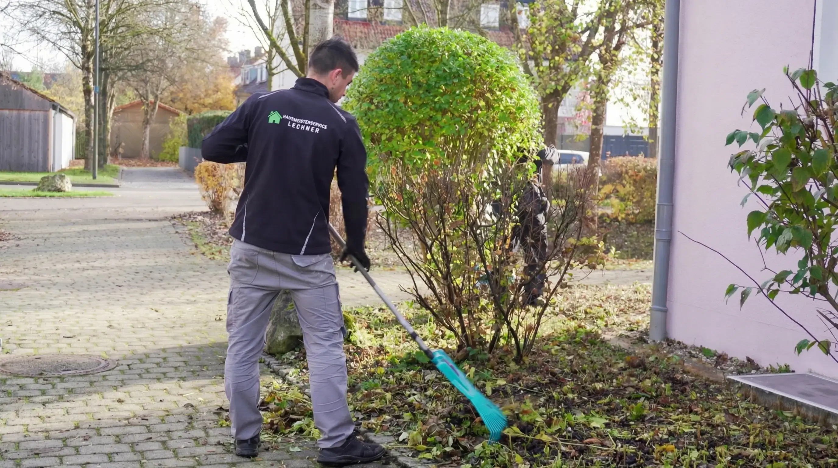 Mann harkt Laub in einer Gartenanlage neben einem Gebäude.