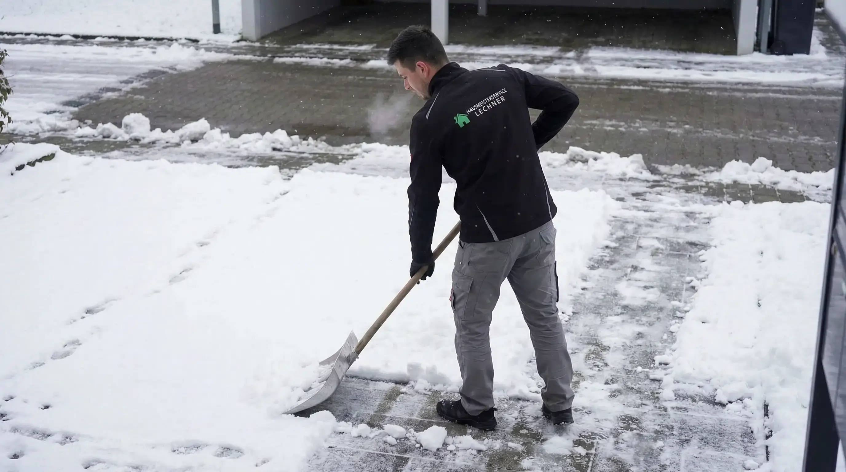 Ein Mann schaufelt Schnee von einer gepflasterten Einfahrt vor einer Garage.