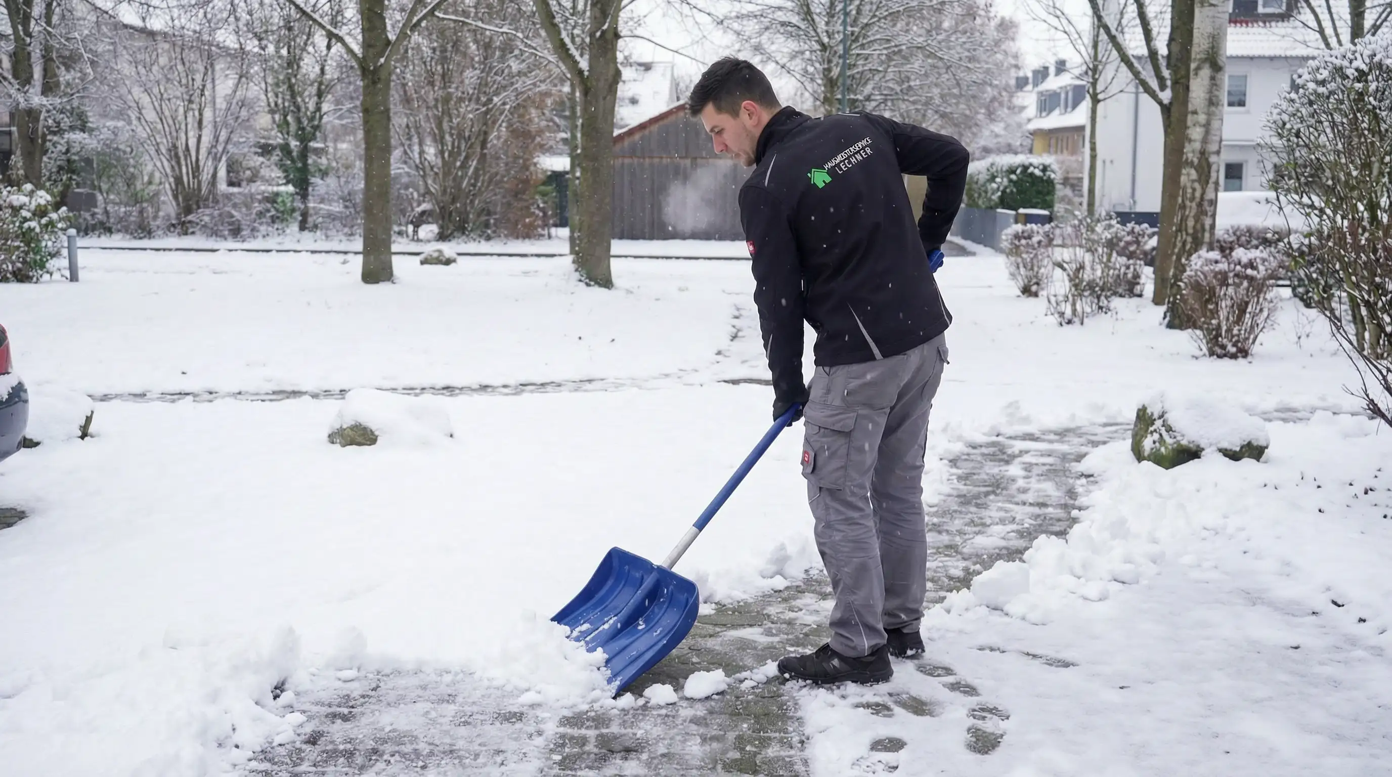 Mann mit blauer Schneeschaufel räumt schneebedeckten Gehweg in einem verschneiten Wohngebiet frei.