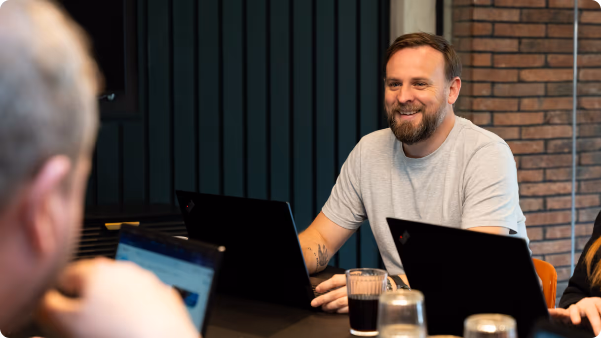 Smiling man with beard sitting at a table with laptops and glasses during a meeting.