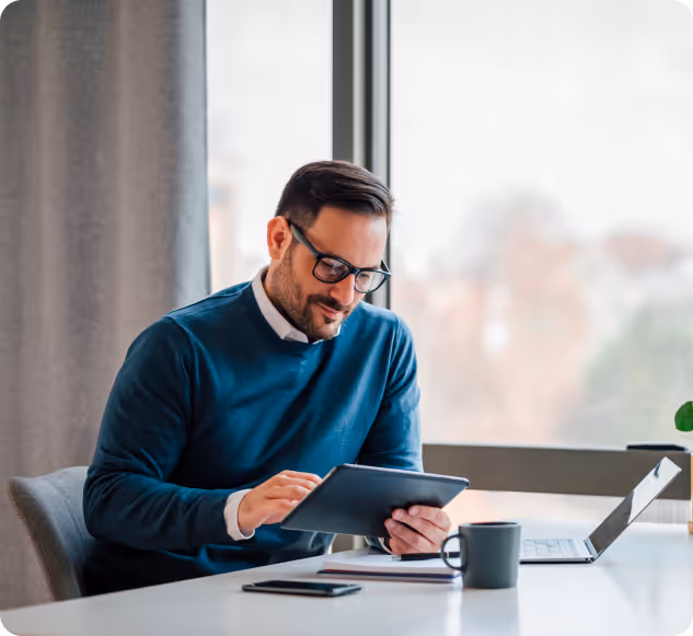 Man wearing glasses and a teal sweater sitting at a desk using a tablet with a laptop, smartphone, and coffee mug nearby.