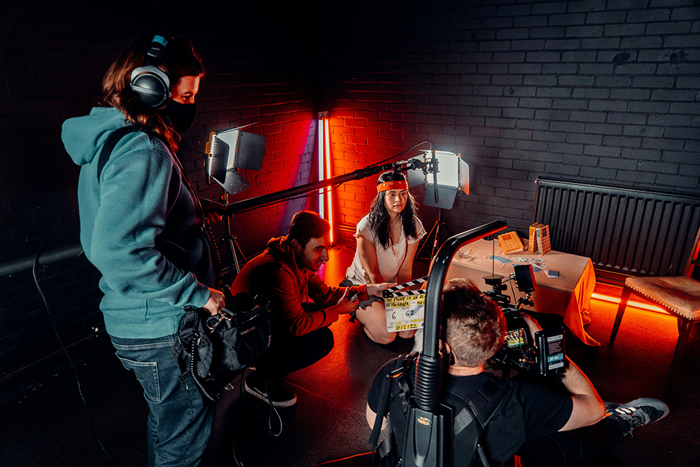 Film crew recording a scene with a female actor holding a clapperboard in a dimly lit room with red lighting and professional video equipment.