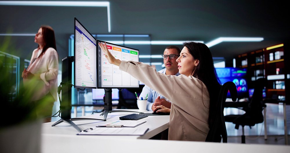 Woman pointing at data on dual monitors while a man watches in a modern office.