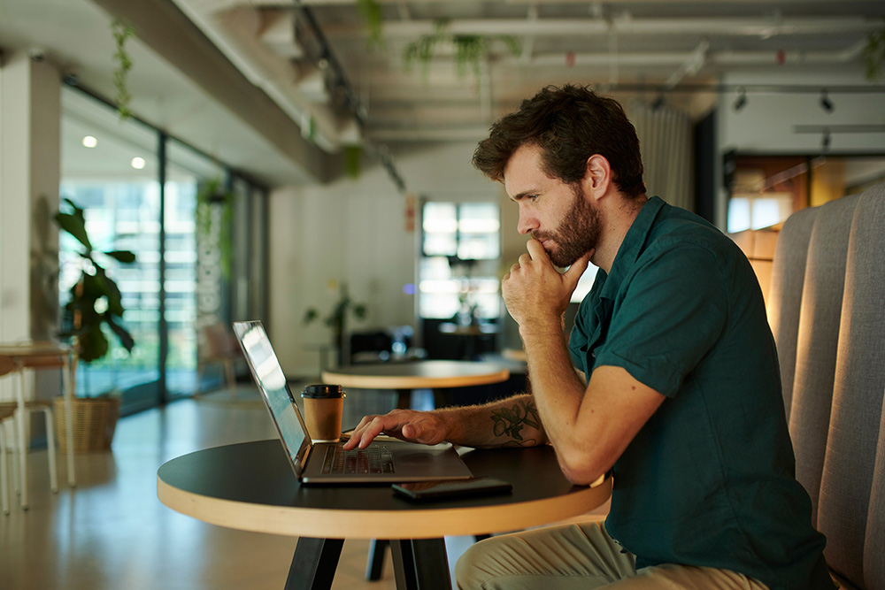Man with beard and tattoo sitting at a round table, working on a laptop in a bright modern office with plants and large windows.