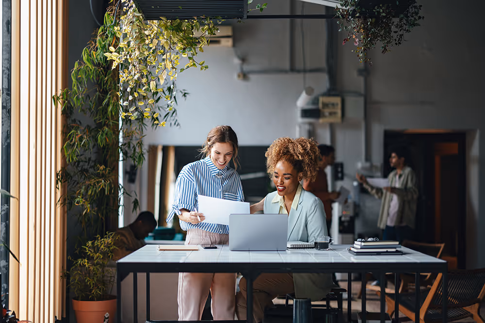 Two women collaborating in an office, one standing and holding a paper while the other sits at a laptop.