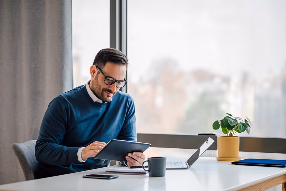 Man with glasses sitting at a white desk using a tablet, with a laptop, coffee mug, and plant nearby.