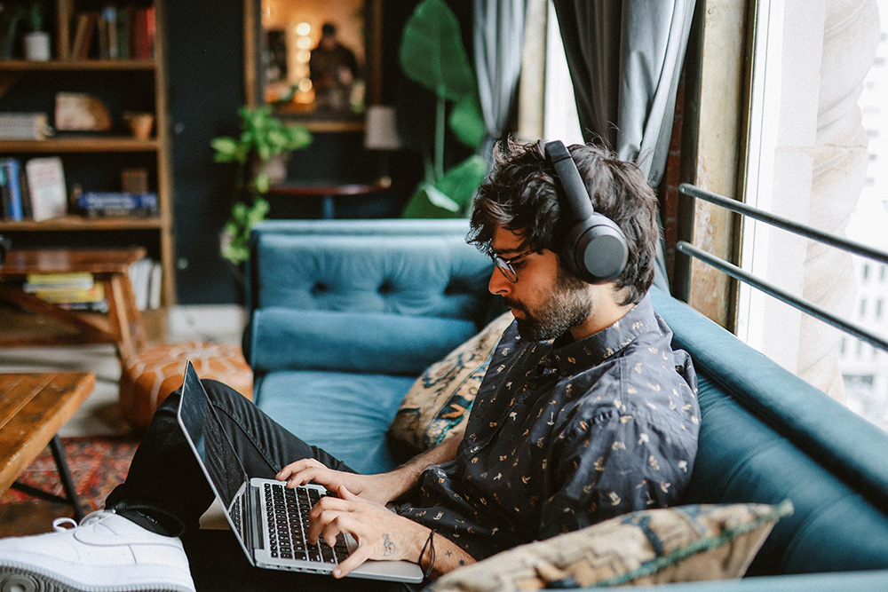 Man with glasses and headphones typing on a laptop while sitting on a blue sofa near a window.