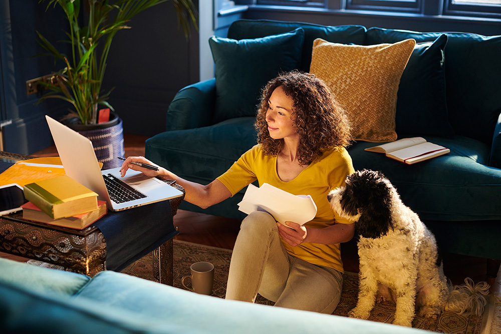 Woman sitting on the floor beside a dog, using a laptop and holding papers in a cozy living room.