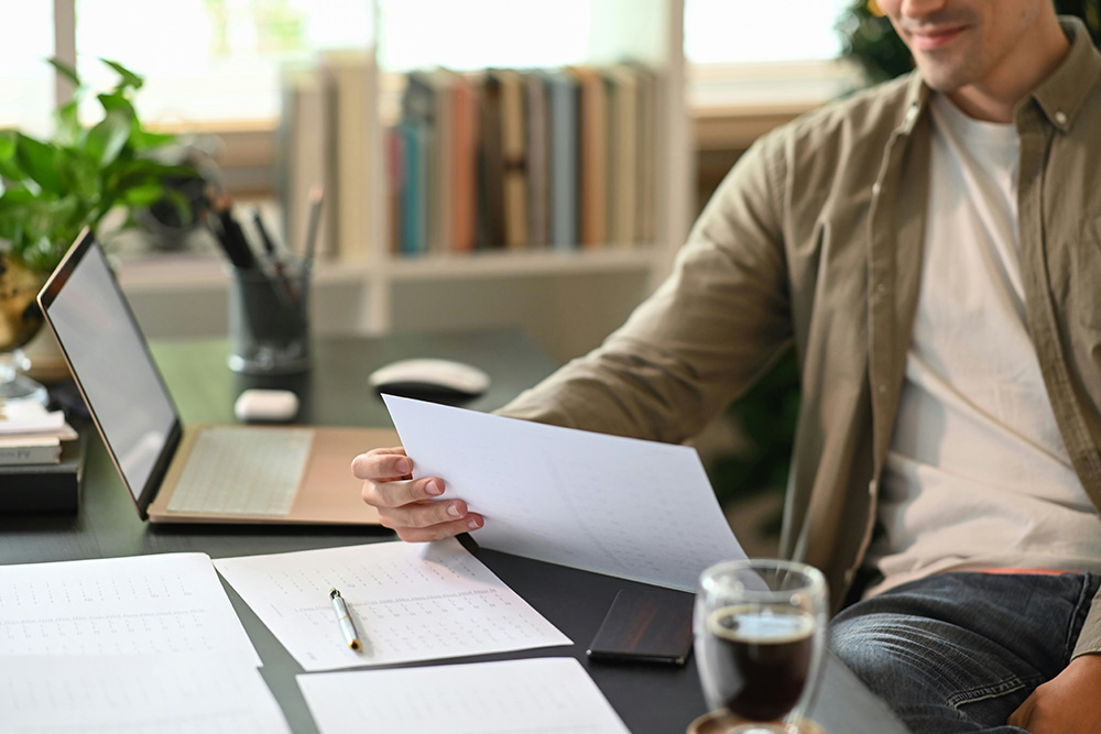 Man sitting at desk holding a sheet of paper with open laptop, documents, and a glass of coffee nearby.