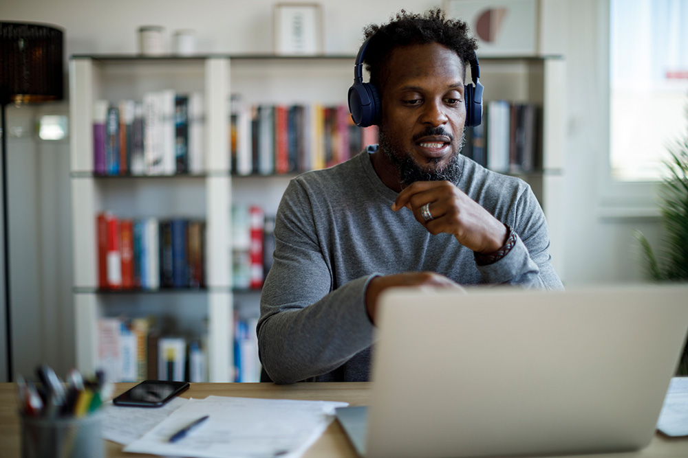 Man wearing headphones and a gray sweater working on a laptop in an office with shelves of books in the background.
