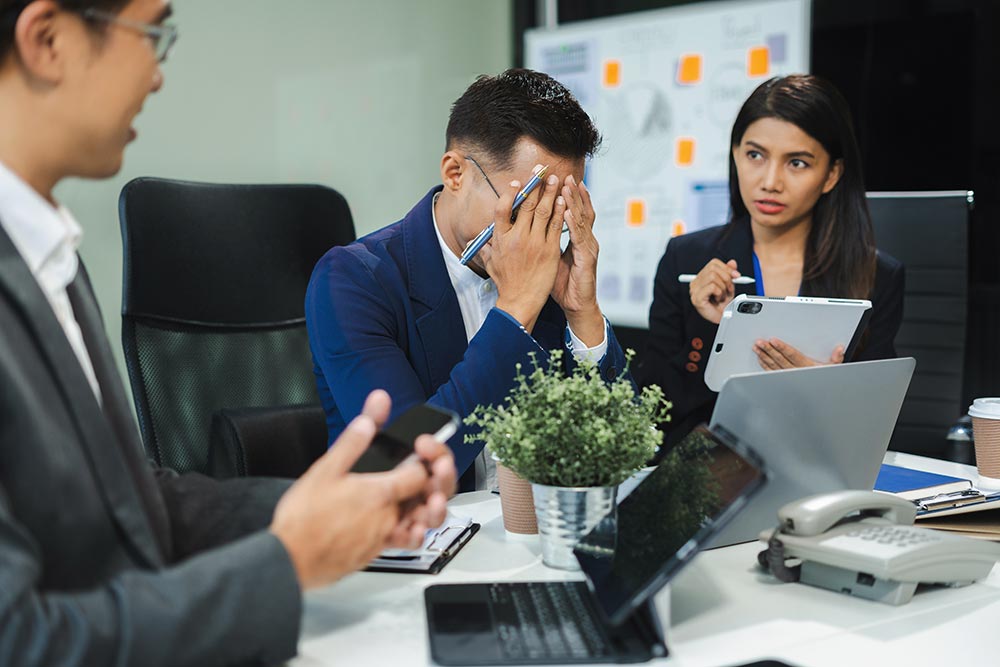 Stressed businessman covering his face while colleagues discuss work in a modern office meeting.