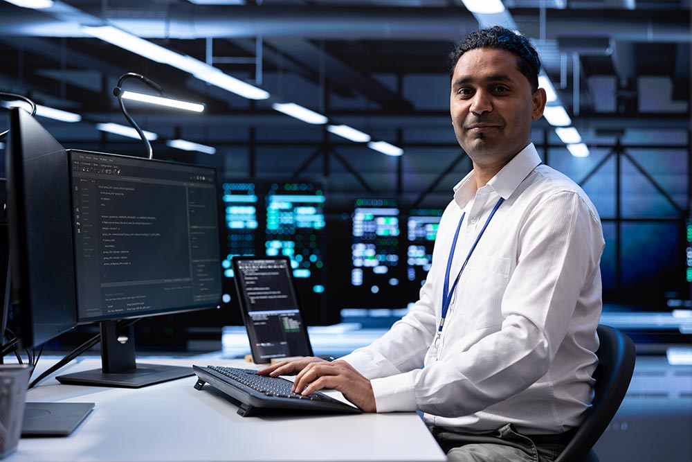 Smiling engineer in a white shirt working at a computer desk with multiple monitors displaying code in a dimly lit control room.
