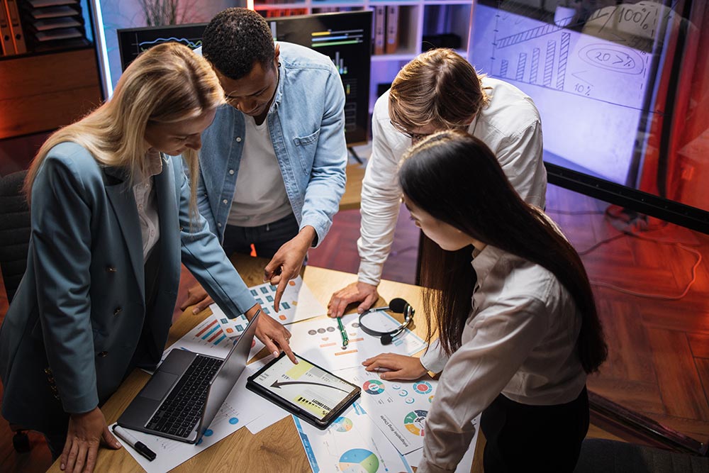 Four business partners gathered around a table reviewing charts and graphs on papers and a tablet in a modern office.