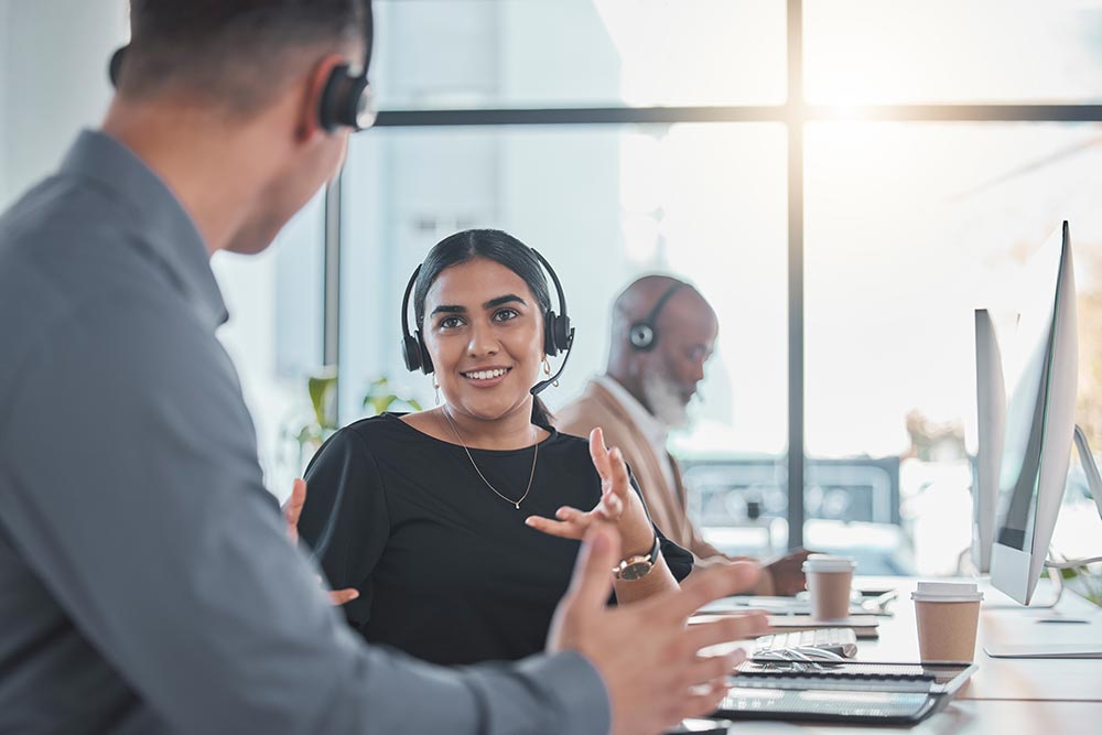 Three call center agents wearing headsets working and having a discussion in a modern office.