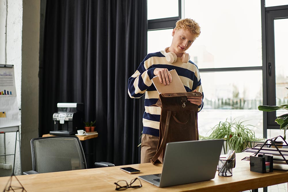 Redhead man in a striped sweater placing a notebook into a brown leather backpack in a modern office.