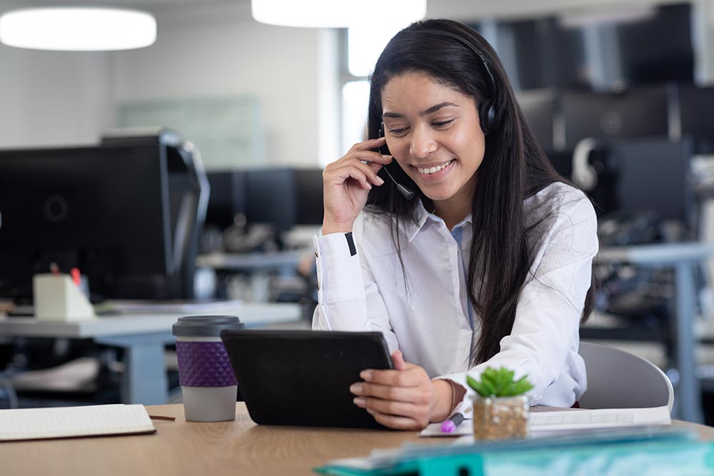Smiling businesswoman wearing a headset and using a digital tablet at her office desk.