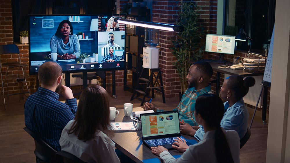 Five people sitting around a table in a brick-walled office engaged in a video conference with four colleagues on a large screen, discussing sales statistics shown on a laptop.