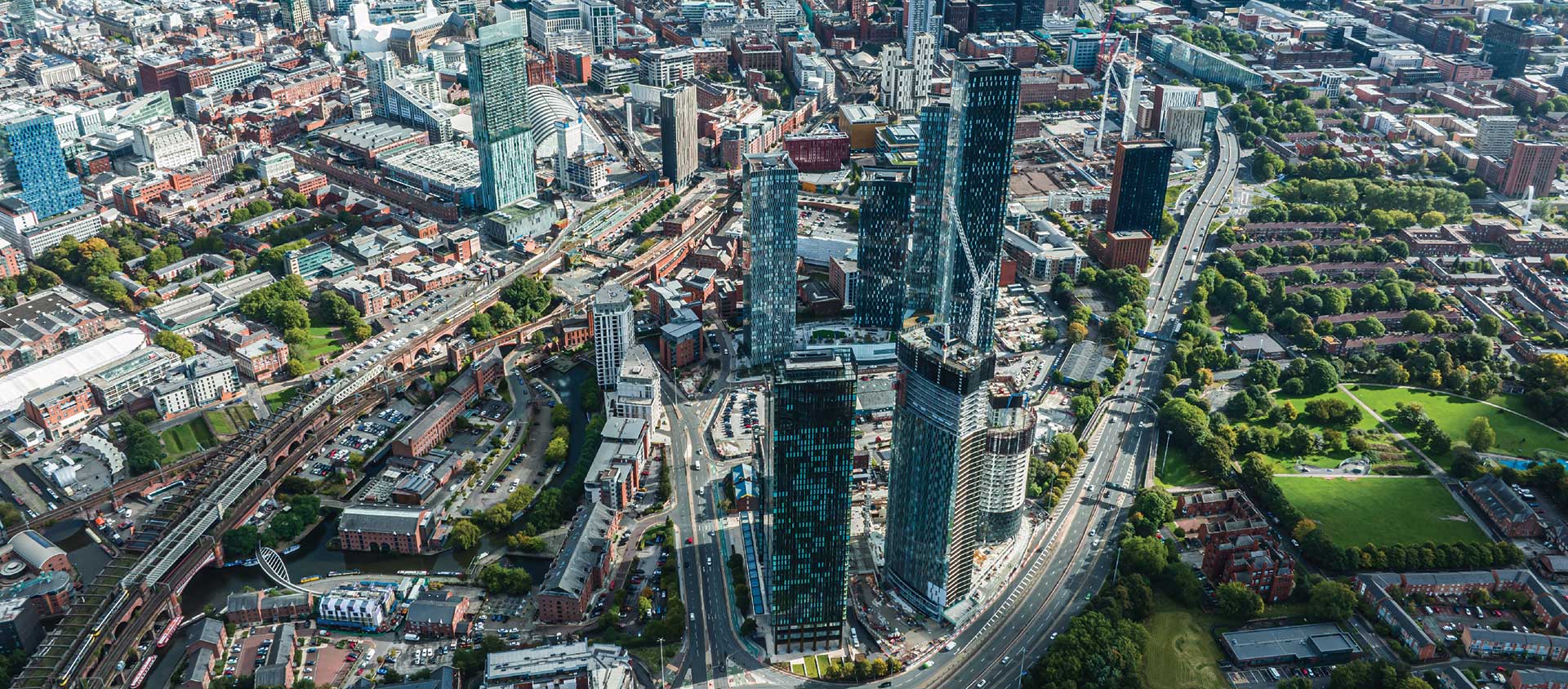 Aerial view of Manchester city center featuring a cluster of modern skyscrapers, surrounding roads, and green park areas.