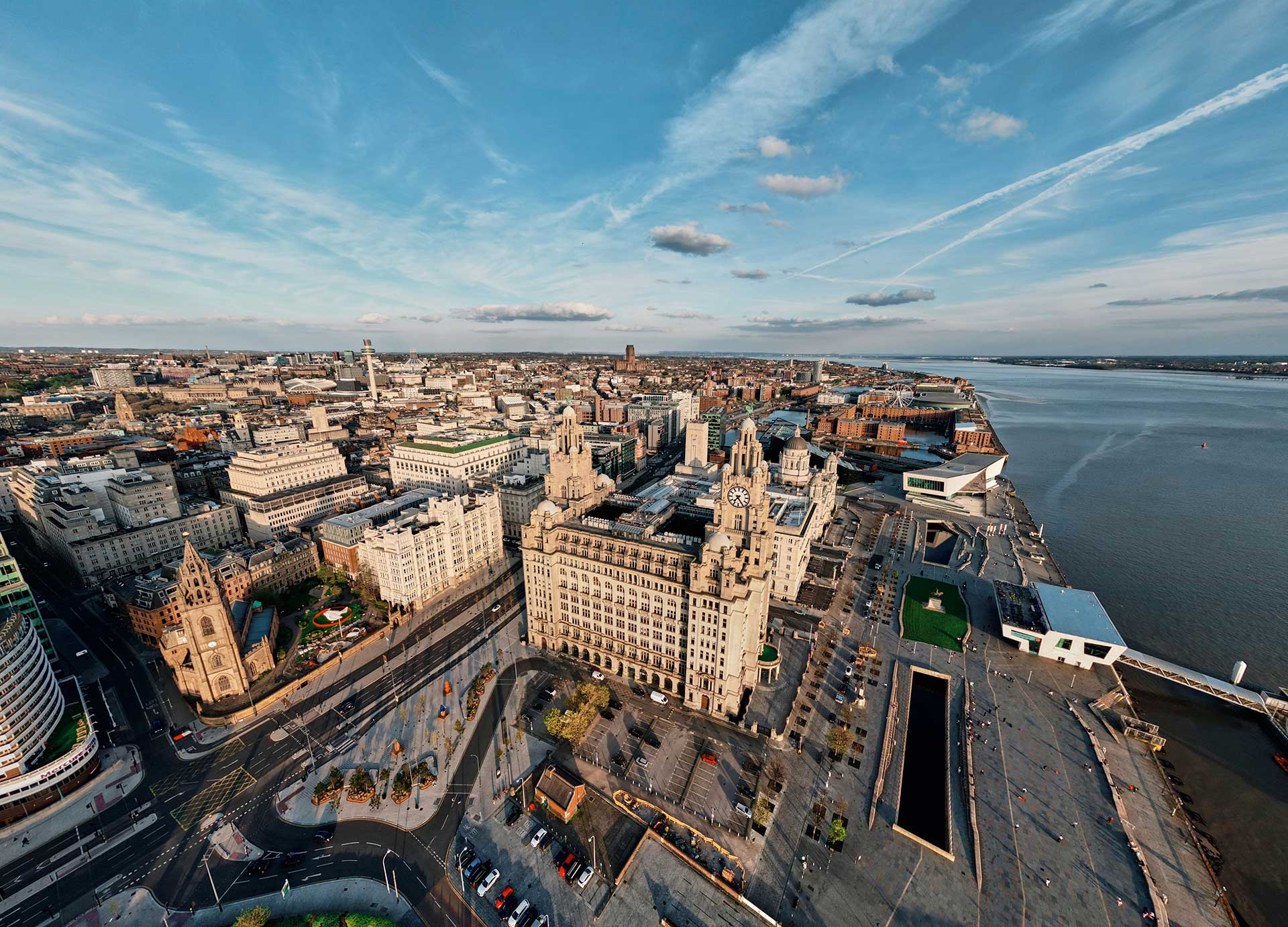 Aerial view of Liverpool waterfront showing the Royal Liver Building, nearby historic buildings, and the River Mersey under a blue sky with scattered clouds.