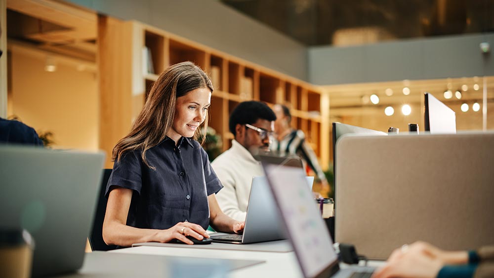 Woman working on a laptop in a modern office with colleagues in the background.
