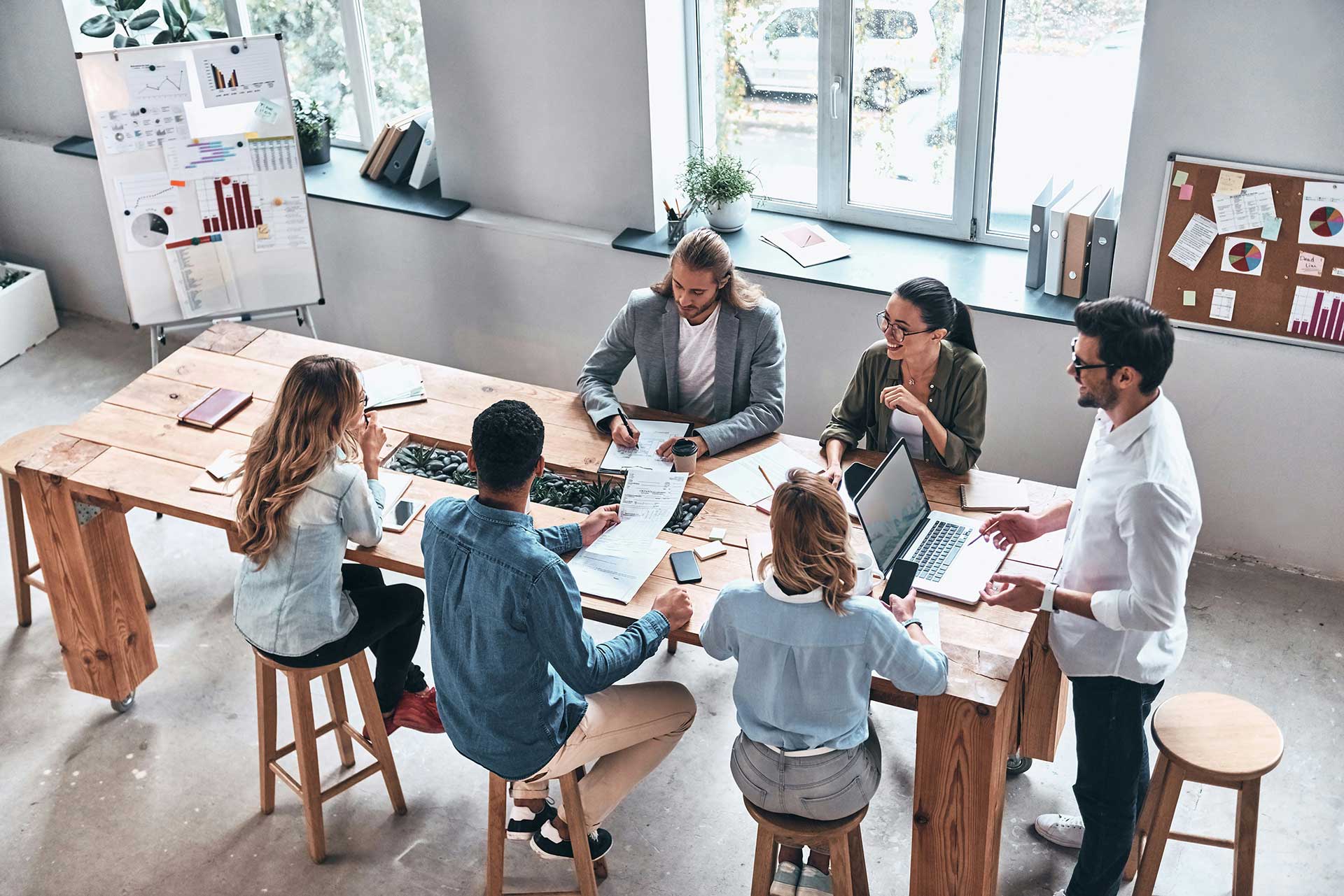 Group of five people collaborating around a wooden table with laptops, documents, and coffee cups in a bright office space.