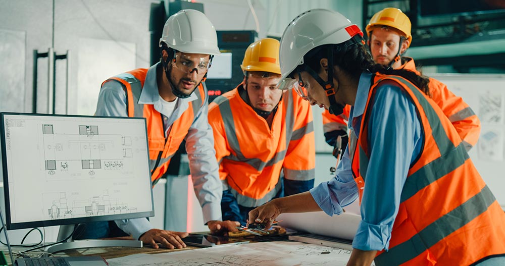 Four engineers wearing safety helmets and orange vests discussing blueprints over a table with a monitor displaying technical drawings.