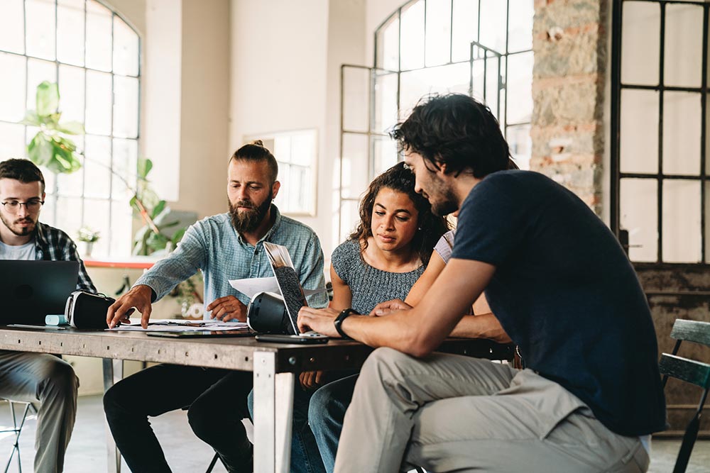 Four young adults collaborating at a wooden table with laptops and documents in a bright, modern workspace.