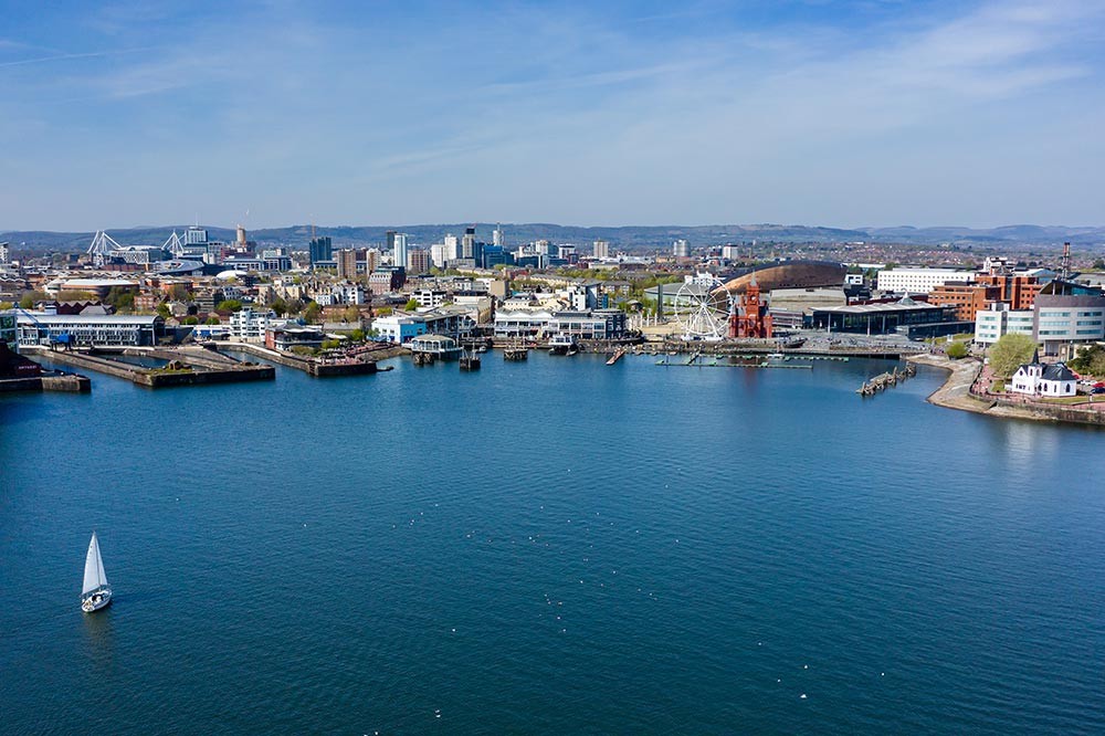 Aerial view of a city waterfront with a sailboat on the water and buildings, including a ferris wheel and modern architecture, under a blue sky.