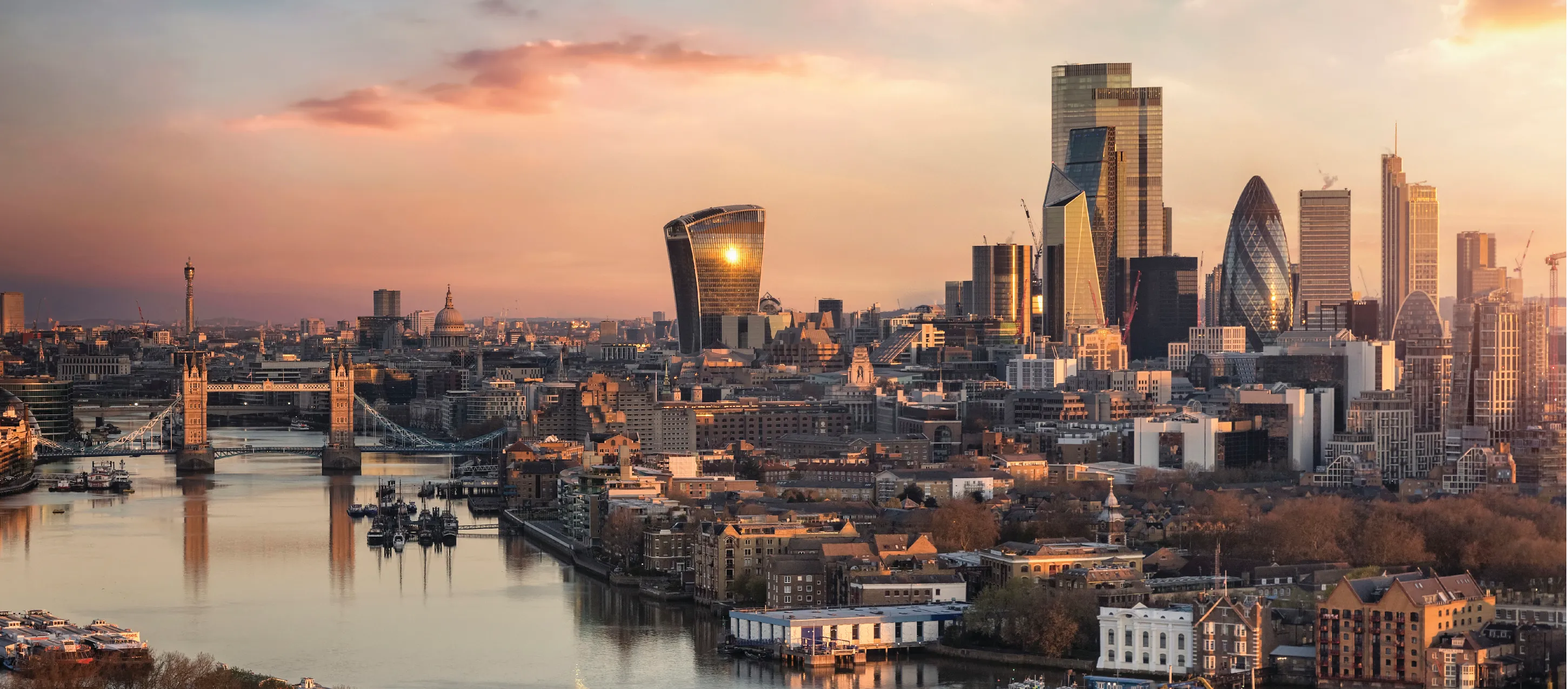 Panoramic view of London at sunset featuring Tower Bridge over the River Thames and the city's modern skyscrapers.