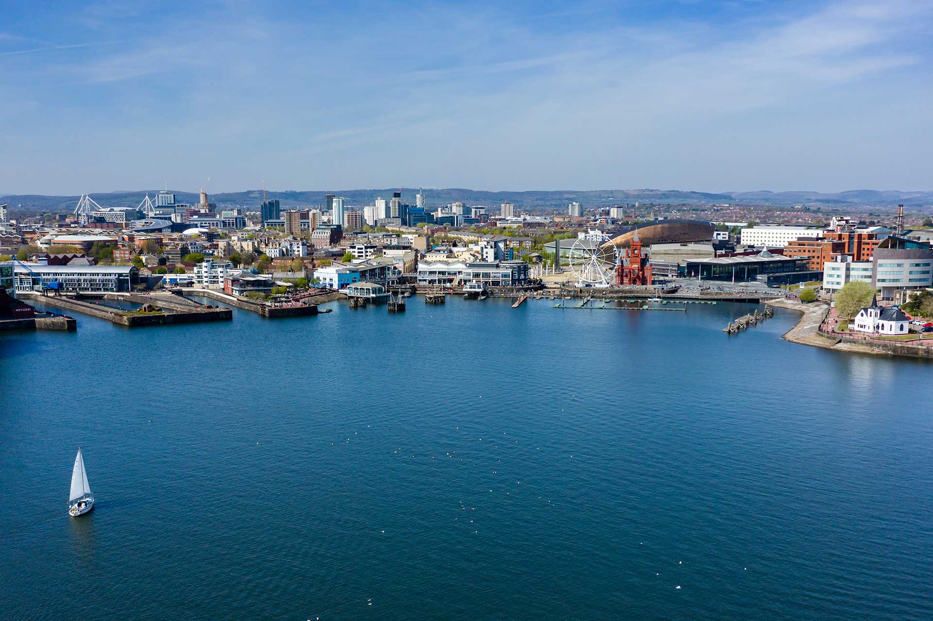 Aerial view of Cardiff Bay with a white sailboat on calm water and the city skyline in the background under a blue sky.