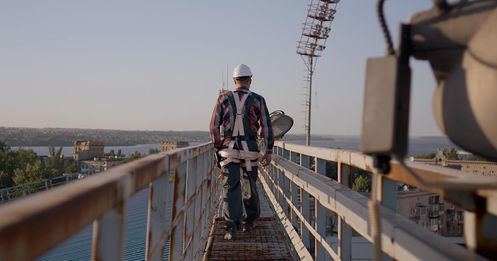 Engineer wearing safety harness and helmet walking on a narrow elevated metal walkway overlooking a cityscape with a river in the background.