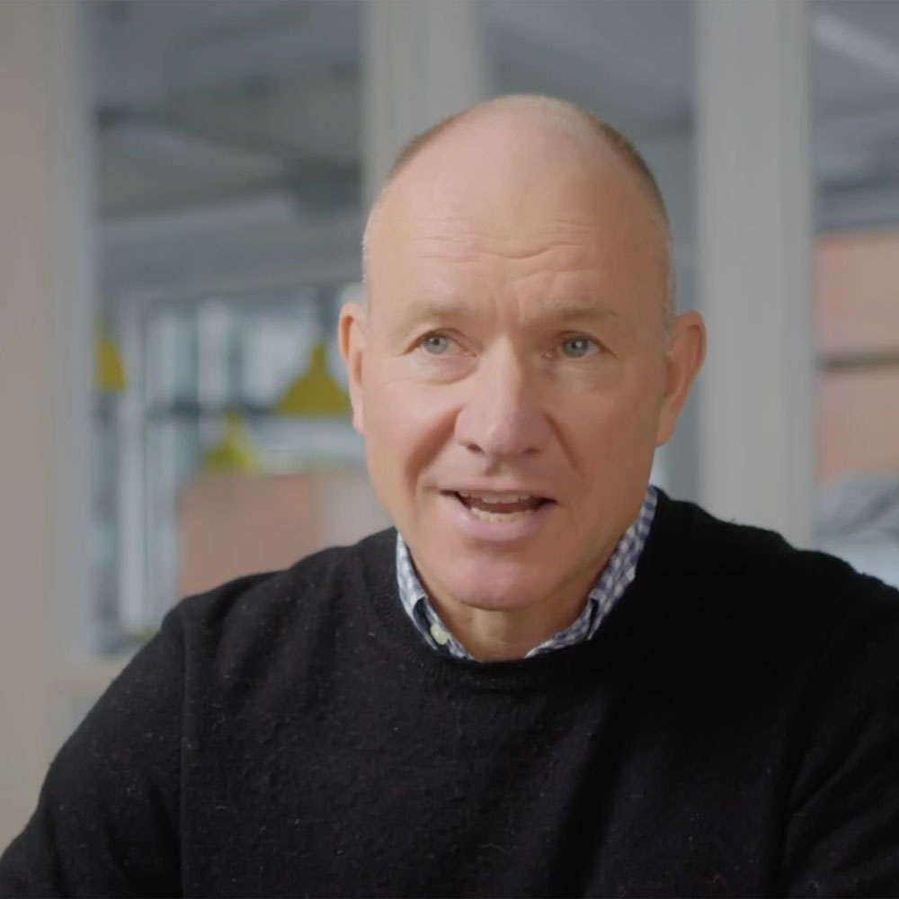 Middle-aged man with short-cropped hair wearing a black sweater over a checkered shirt, speaking indoors with blurred windows and yellow hanging lights in the background.