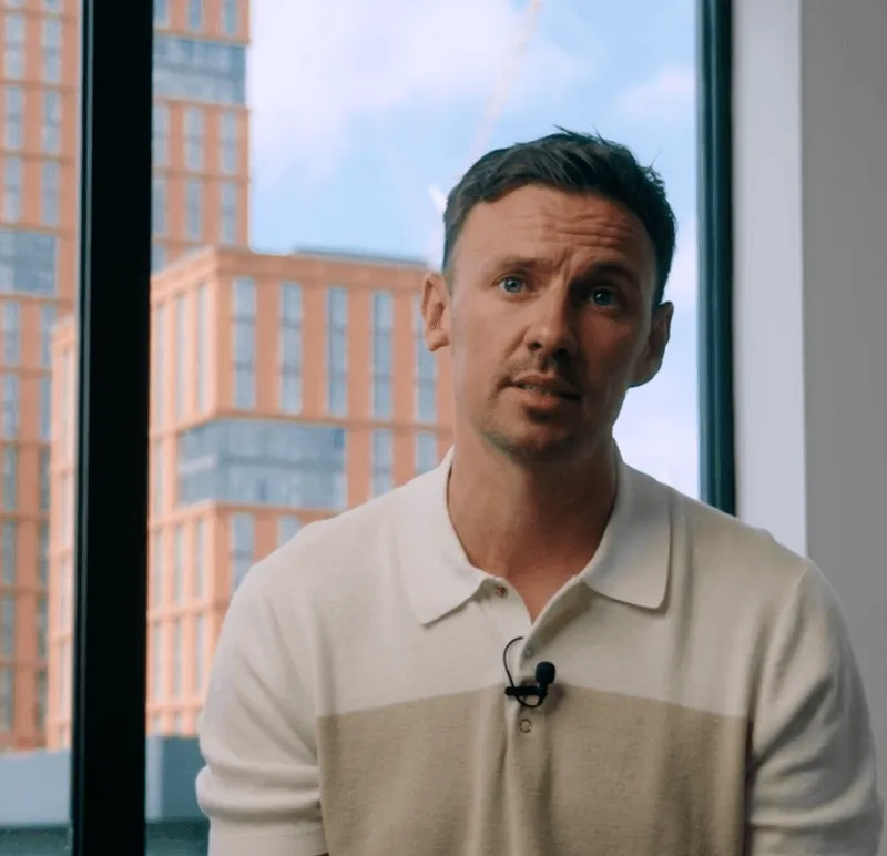 Man wearing a white and beige polo shirt with a microphone clipped, sitting indoors in front of large windows showing a blurry cityscape with modern buildings.