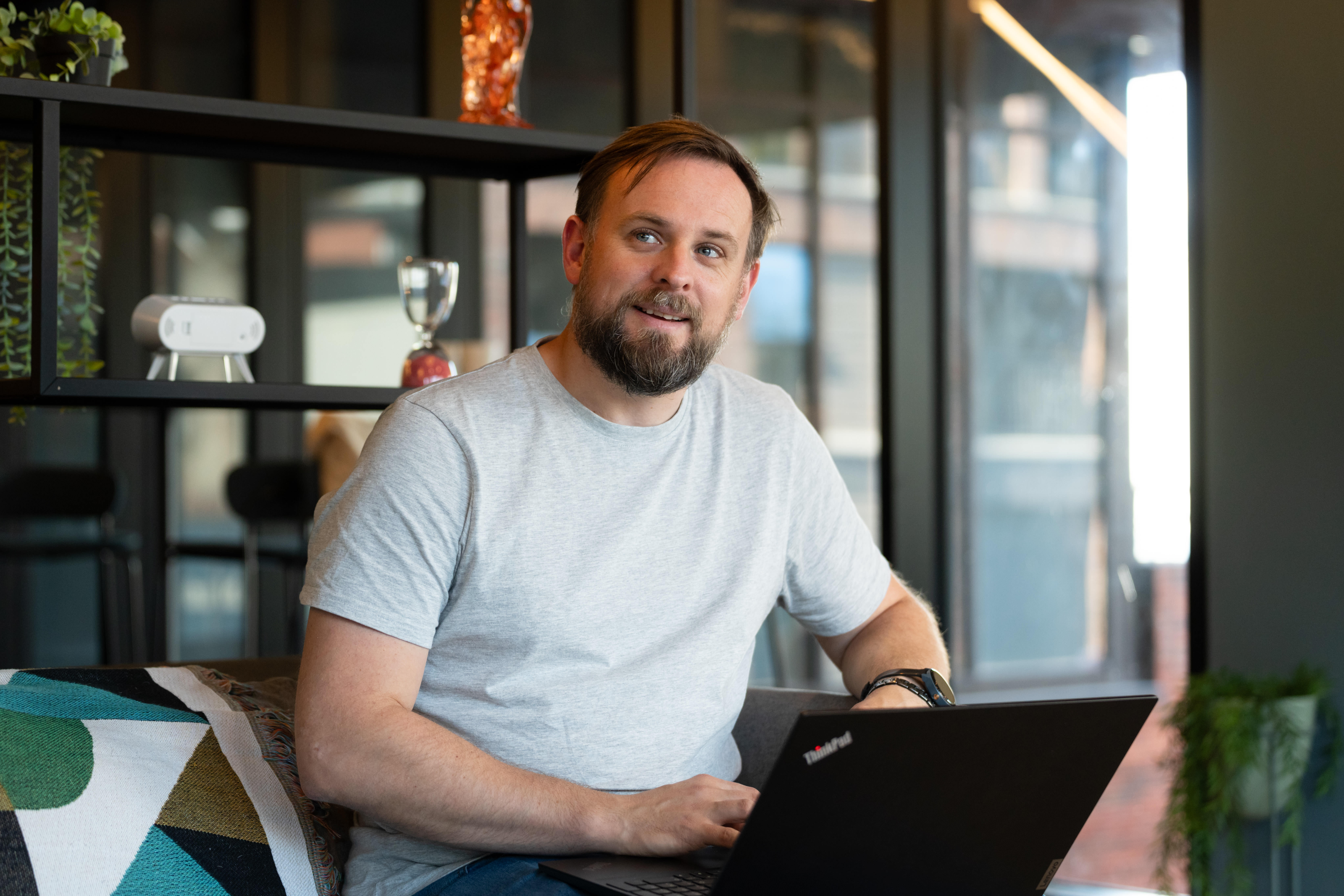 Man with beard in a gray t-shirt sitting on a couch, using a black ThinkPad laptop in a modern office setting.
