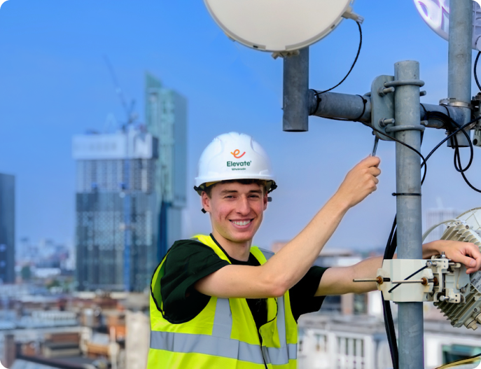 Smiling technician in a white hard hat and yellow safety vest working on telecommunications equipment on a rooftop with city buildings in the background.