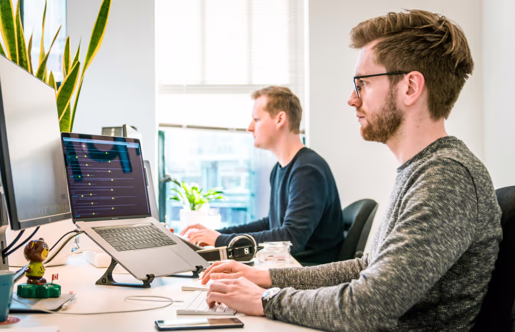 Two men working at computers in a bright office with plants and multiple screens.