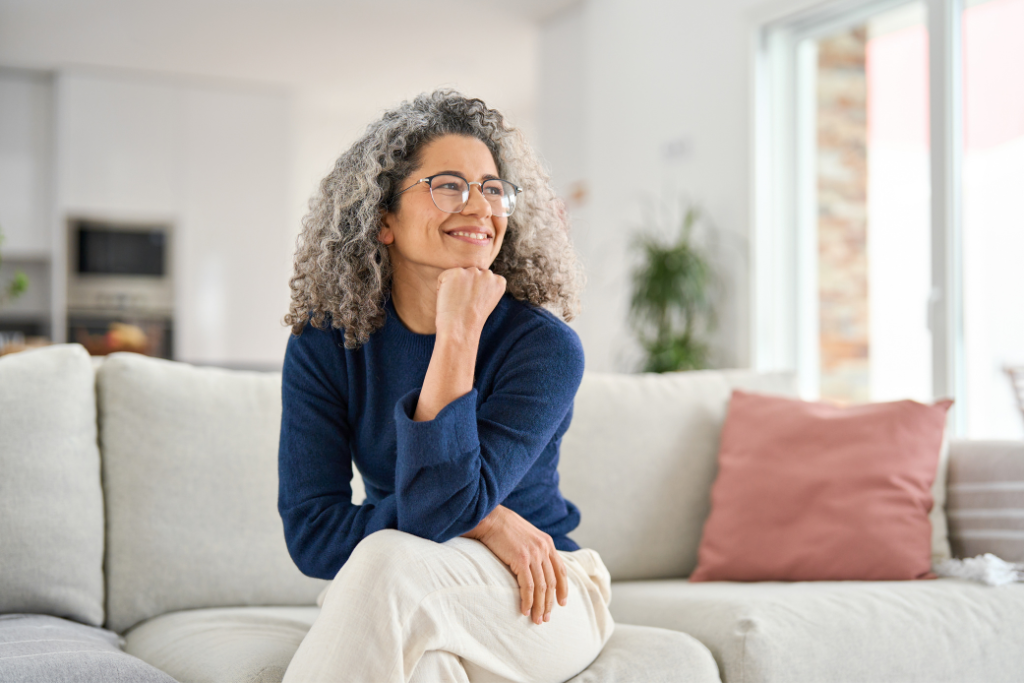 Smiling middle-aged woman with curly gray hair and glasses sitting on a light gray couch in a bright living room.