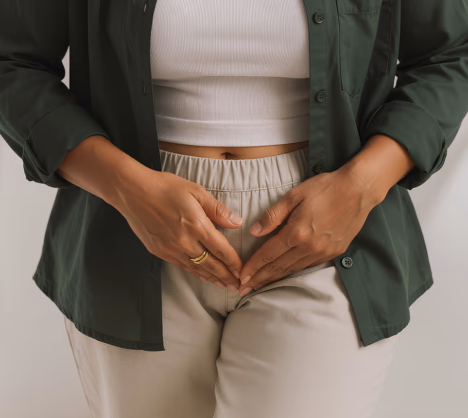 Woman wearing beige pants and a green shirt forming a heart shape with her hands over her lower abdomen.
