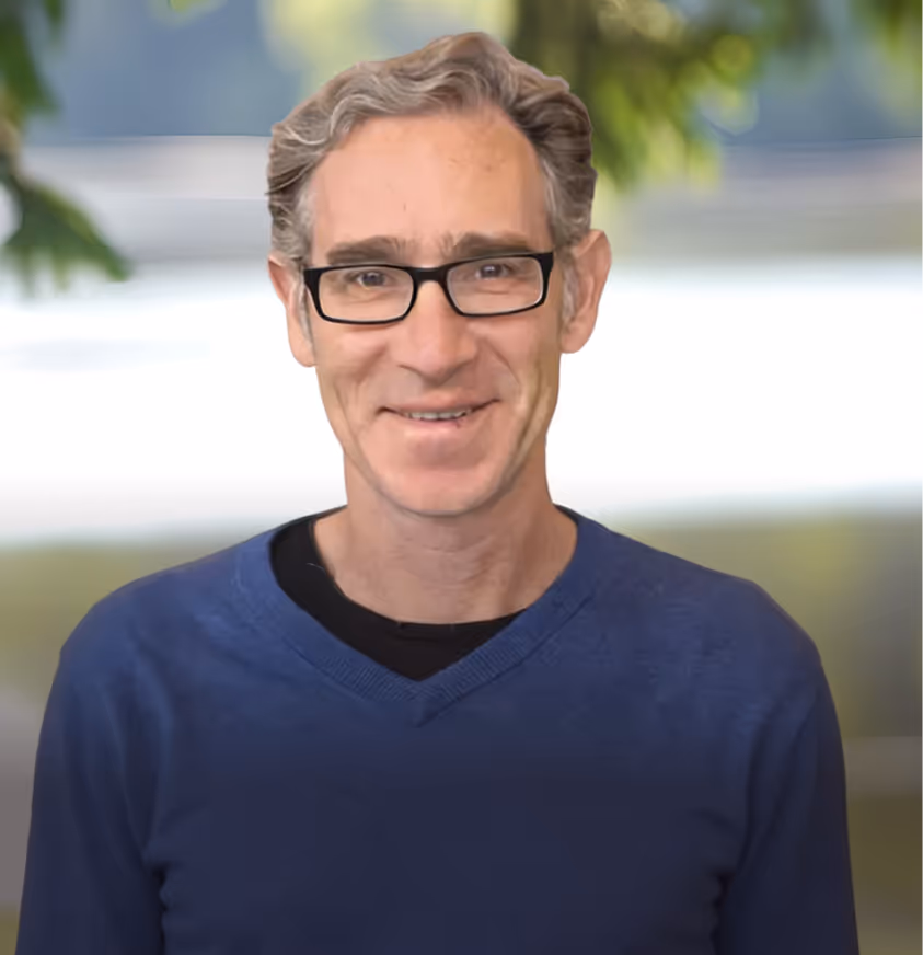 Middle-aged man with glasses and gray hair wearing a blue V-neck sweater smiling in front of a blurred outdoor background.
