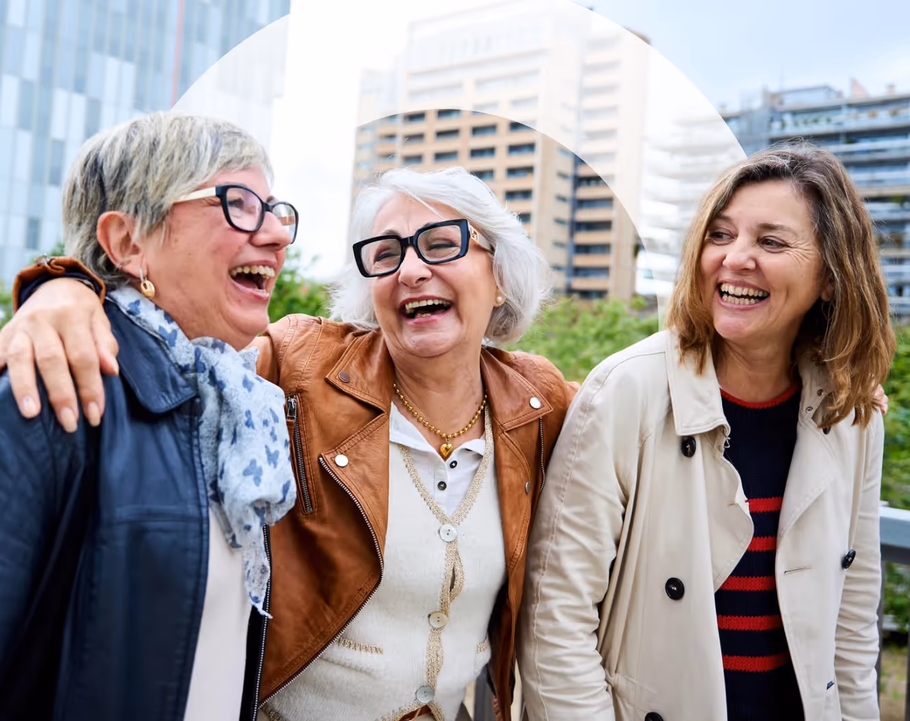 Three middle-aged women wearing jackets, laughing together outdoors with urban buildings in the background.