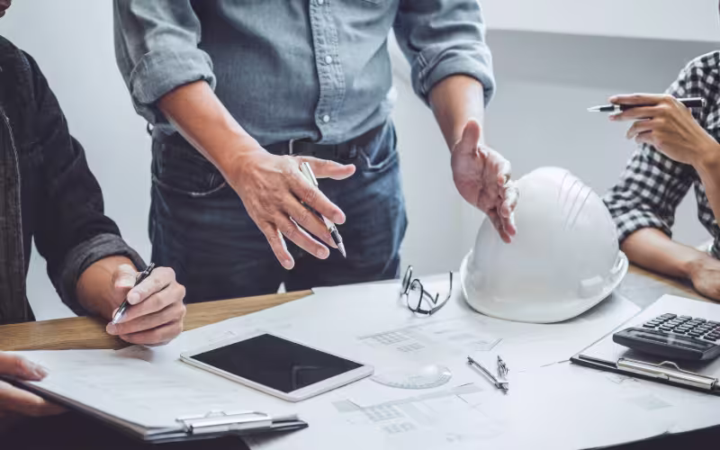Three people sitting around a desk looking at drawings. There is also a hard hat on the desk along with pens, a tablet and a calculator. 