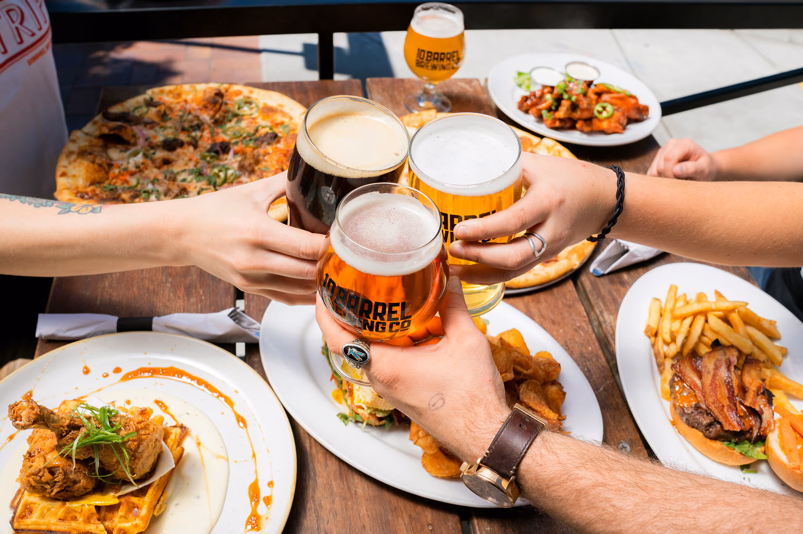 Four hands clinking beer glasses over table of food