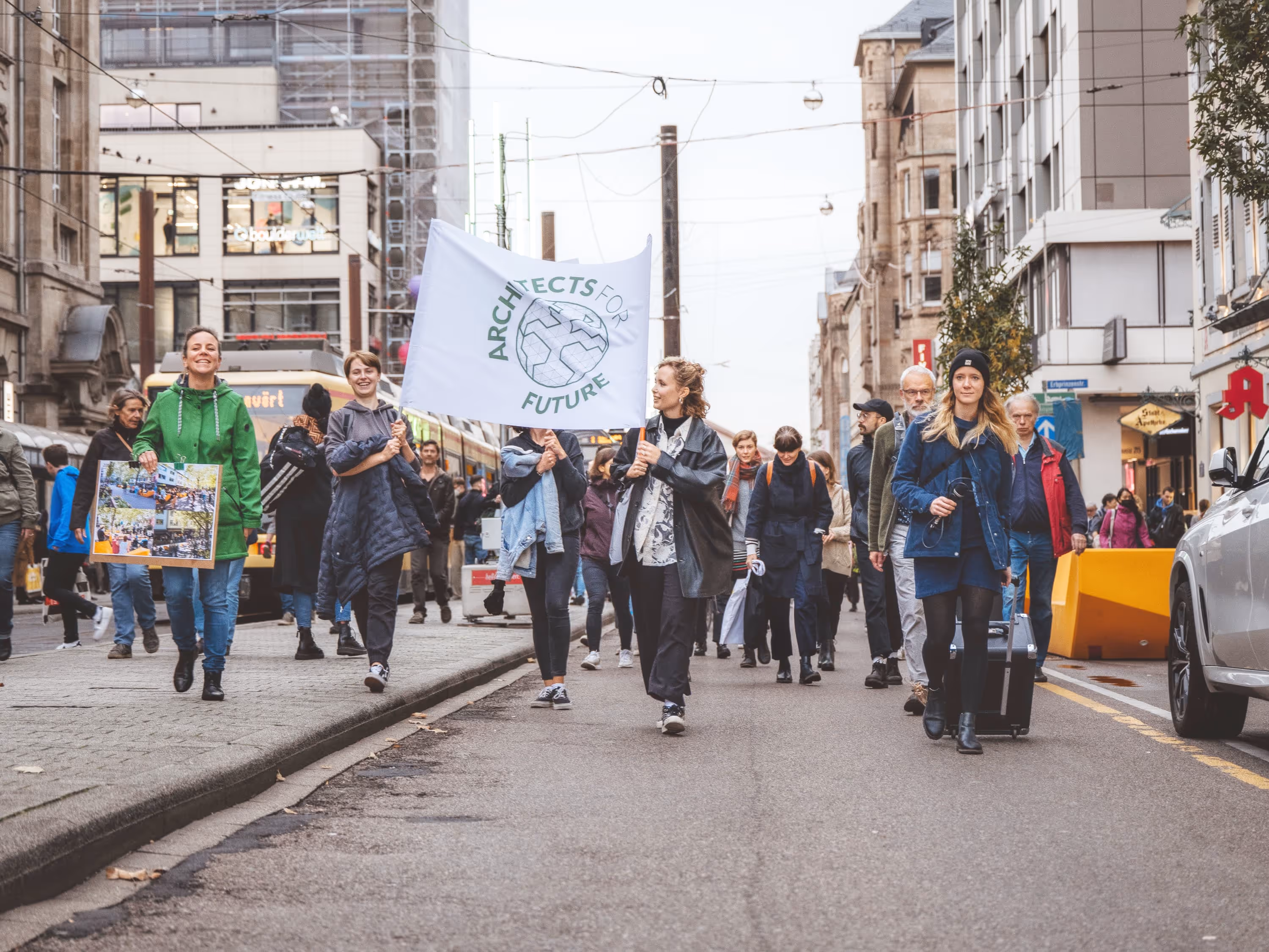 Menschen marschieren auf einer Straße in der Stadt, eine Person hält eine Flagge mit der Aufschrift ‚Architects for Future‘, eine andere trägt ein Plakat.