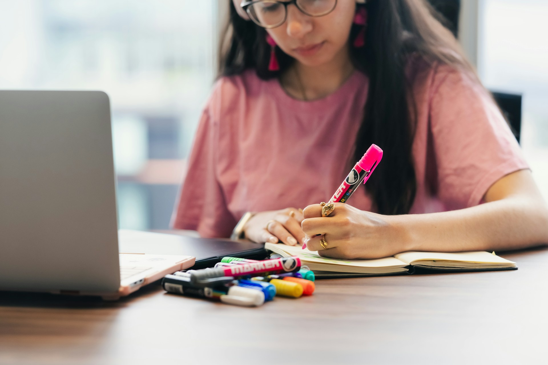 girl with a pinkk shirt writing by hand on a desk with a laptop open in front of her
