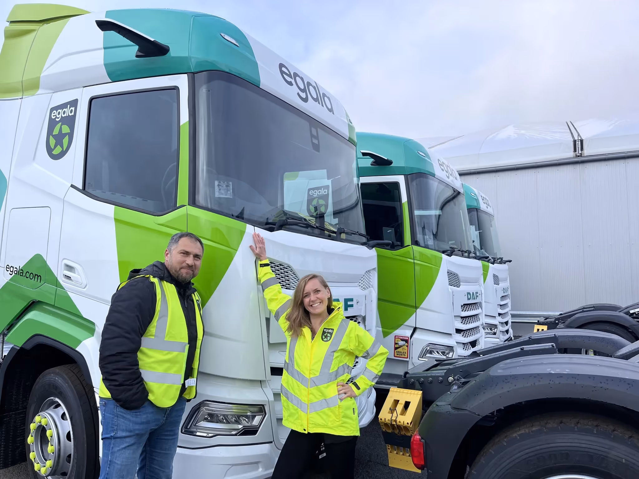 Two people in high-visibility vests smiling and posing in front of Egala by Boekestijn trucks.